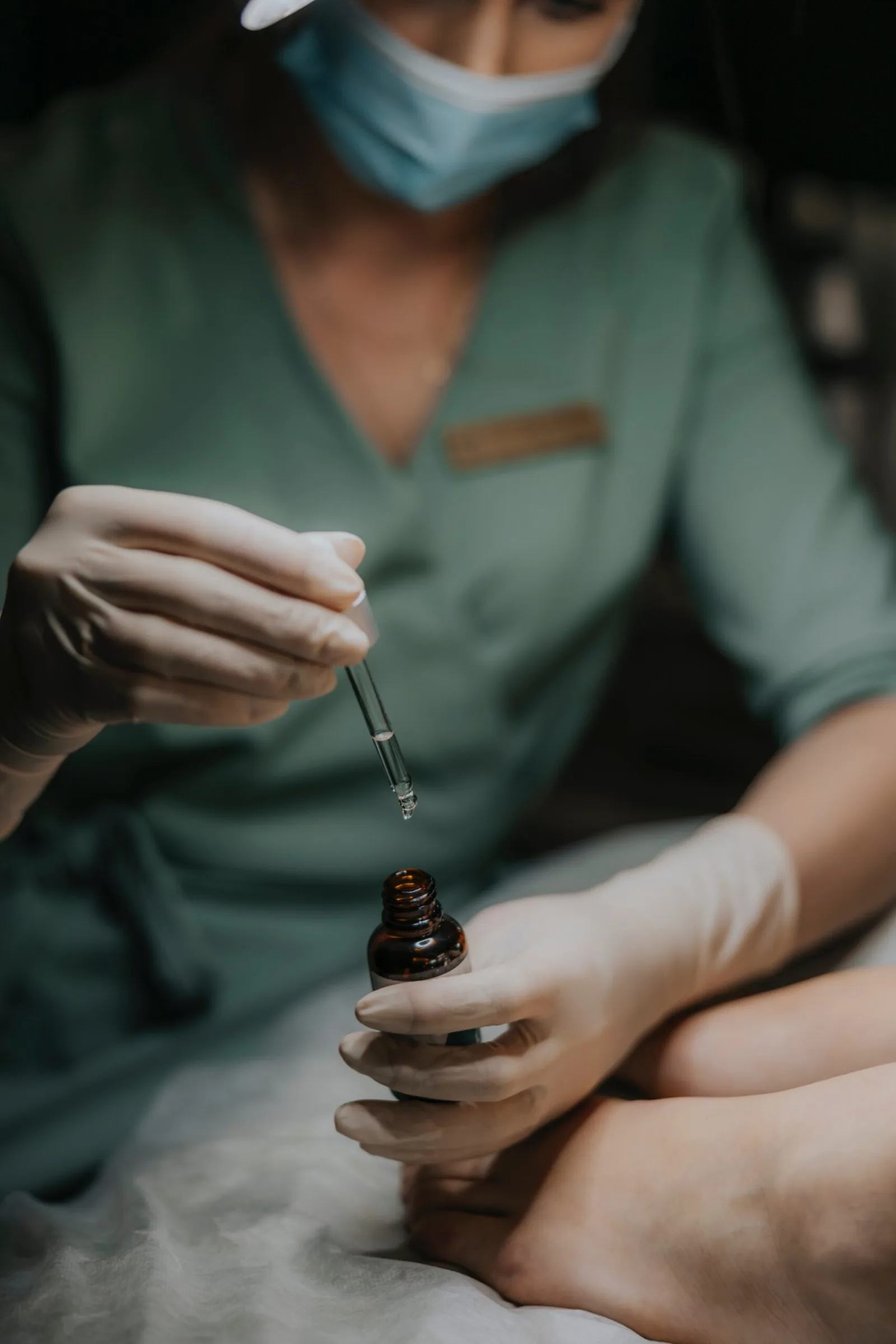 Kosmetolog w zielonym uniformie i maseczce nabiera pipetą preparat z ciemnej buteleczki nad stopą pacjentki