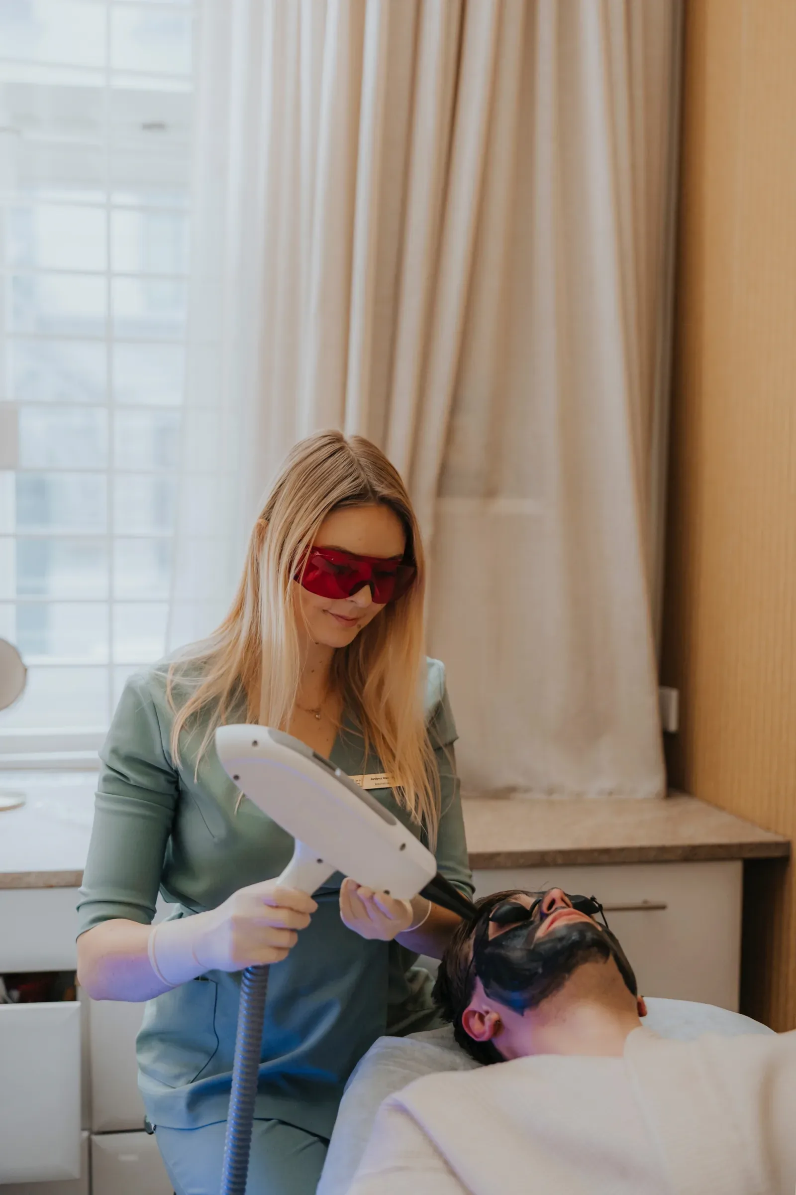 Blond kosmetolozka w mietowym uniformie i czerwonych okularach ochronnych prowadzi glowice lasera nad twarza lezacego me