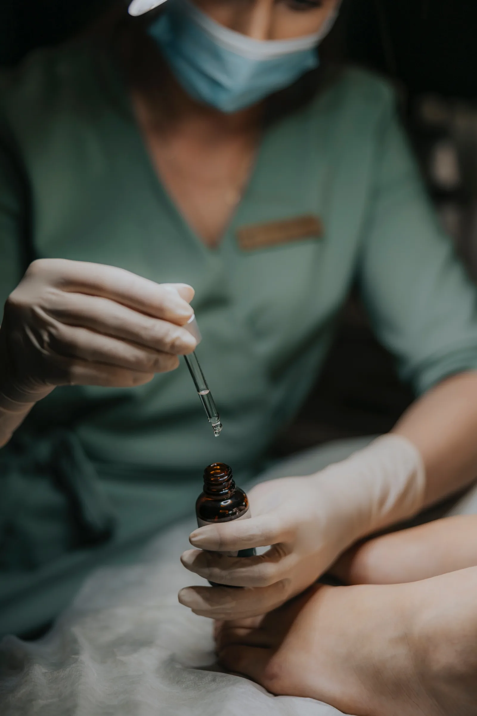 Kosmetolog w zielonym uniformie i maseczce nabiera pipetą preparat z ciemnej buteleczki nad stopą pacjentki